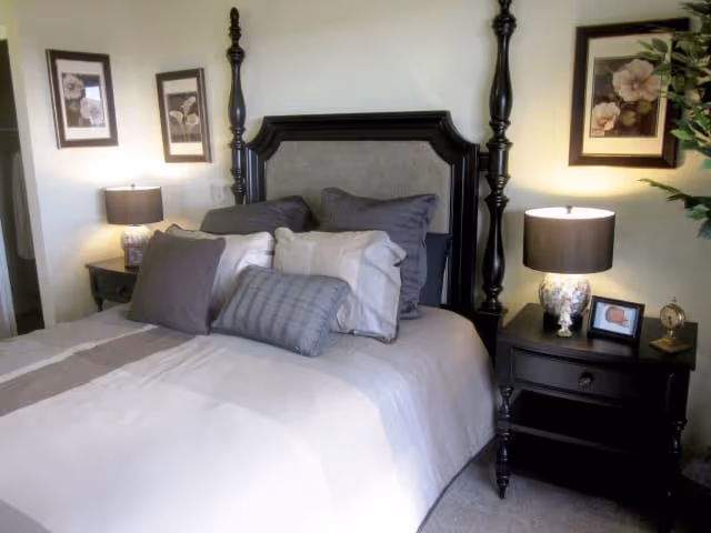 A neatly made bed with multiple pillows in shades of gray and white, flanked by two black nightstands each with a lamp and decorative items. The bed has a dark wooden headboard with tall posts. Two framed floral pictures hang on the wall above the nightstands.