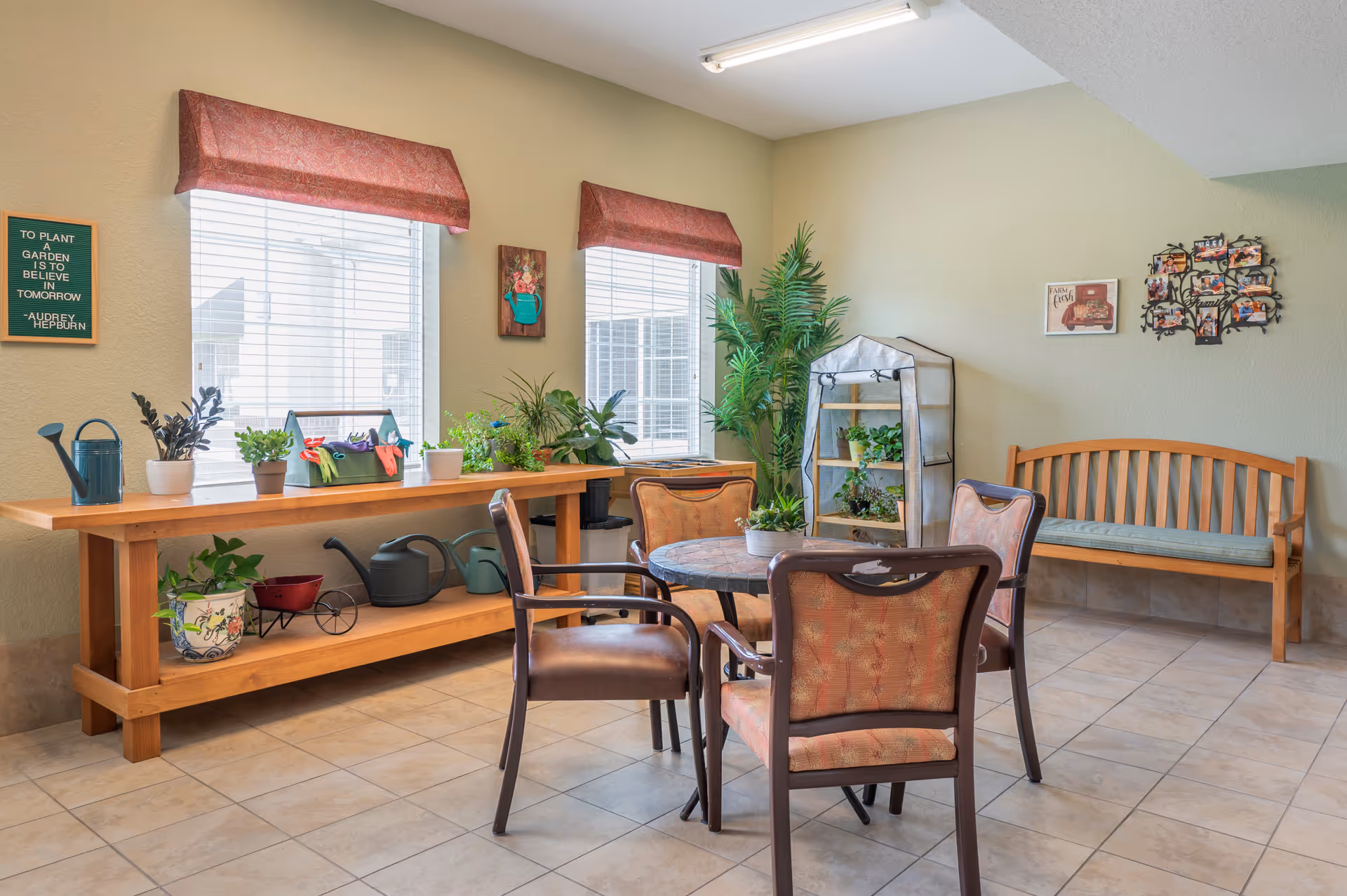 A bright indoor sitting area with a round table surrounded by four cushioned chairs. Behind the table is a wooden bench with a cushion. To the left, a wooden console table holds various potted plants, gardening tools, and watering cans. Two windows with red valances let in natural light. The walls are decorated with framed pictures and a quote board.