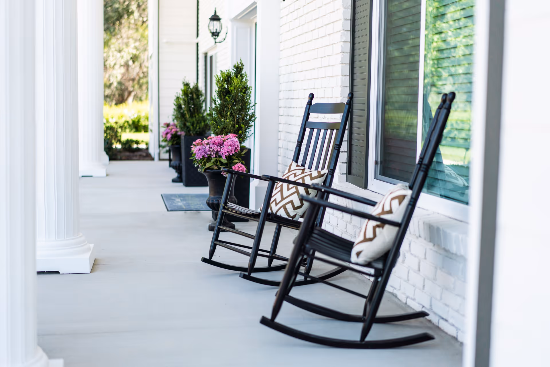 A porch area with two black rocking chairs each with a patterned cushion, white brick wall with windows, white columns, and potted plants with pink flowers and greenery.