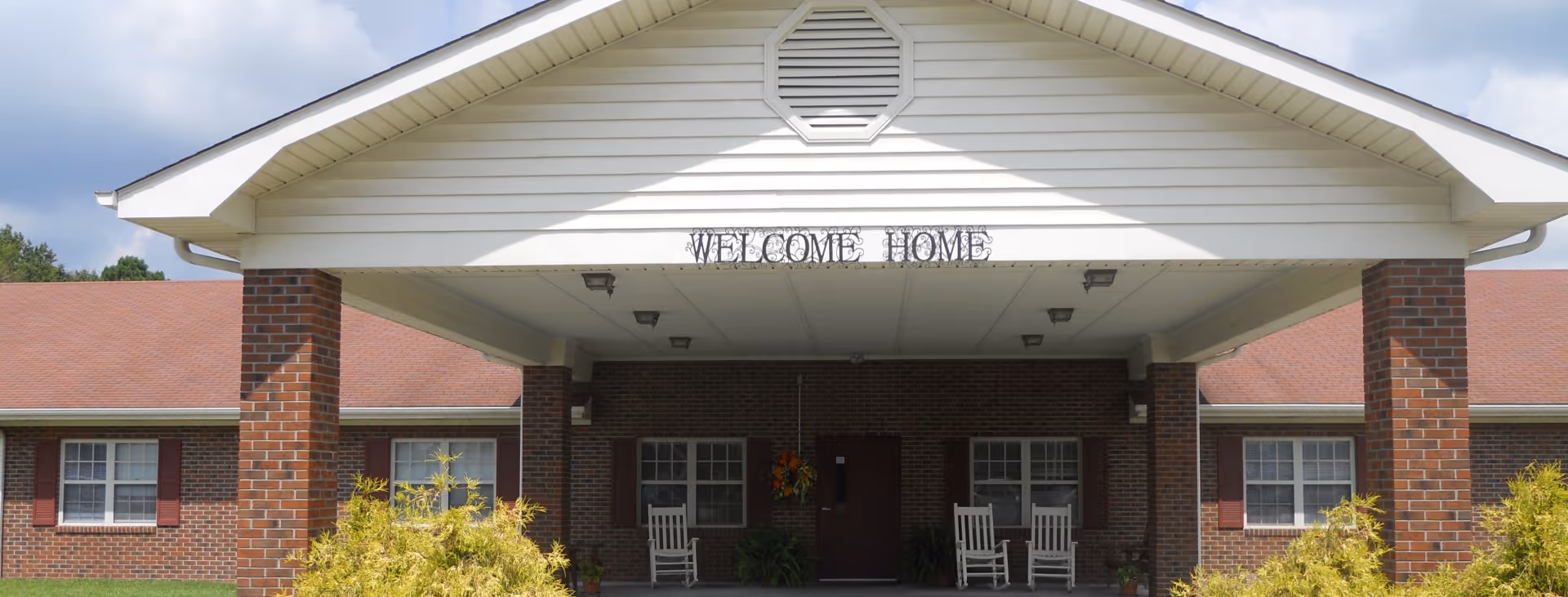 Front exterior view of a brick building with a covered entrance supported by brick pillars. The entrance has a white gabled roof with the words 'WELCOME HOME' displayed above. There are white rocking chairs on the porch and some greenery in front of the building.