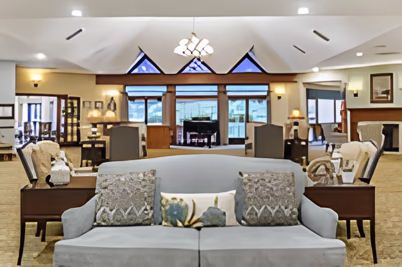 Spacious and well-lit living room area in a senior living facility with a light gray sofa adorned with three decorative pillows in the foreground. The room features multiple seating arrangements including armchairs and side tables with lamps. Large windows and triangular transom windows allow natural light to fill the space. A chandelier hangs from the ceiling, and a fireplace is visible on the right side of the room.