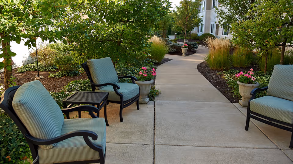 Outdoor courtyard walkway with cushioned chairs, potted flowers, and landscaped greenery at a senior living facility.