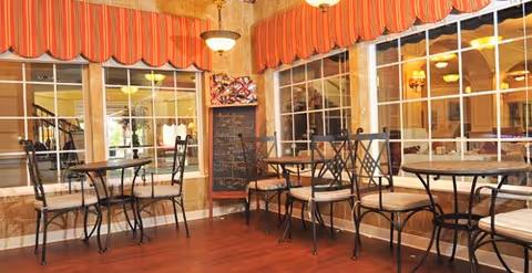 A cozy dining area with several small round tables and metal chairs with cushions. The room features large windows with red and orange striped valances, warm lighting fixtures, and a chalkboard menu on the wall. The floor is wooden, and the overall ambiance is inviting and comfortable.