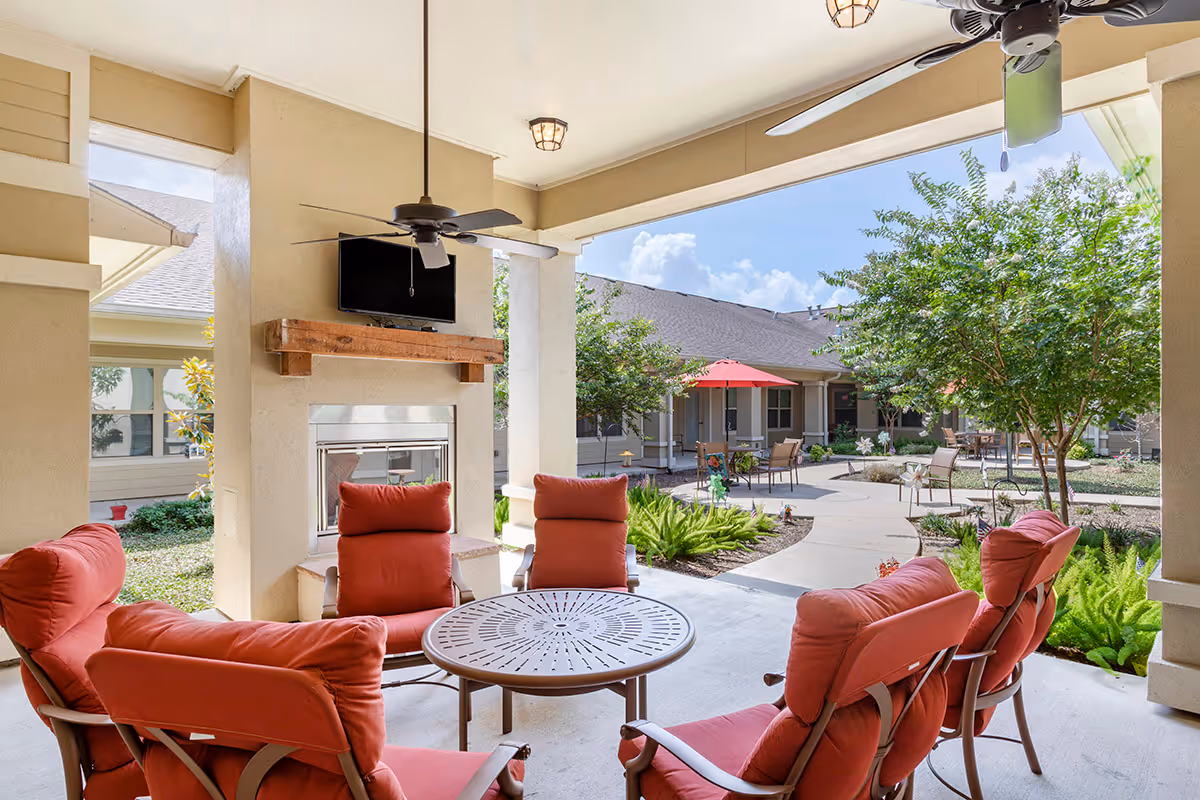 Covered outdoor seating area with six cushioned chairs arranged around a round metal table. The area features a ceiling fan, a mounted TV above a fireplace, and overlooks a garden courtyard with trees, plants, and additional seating with umbrellas.