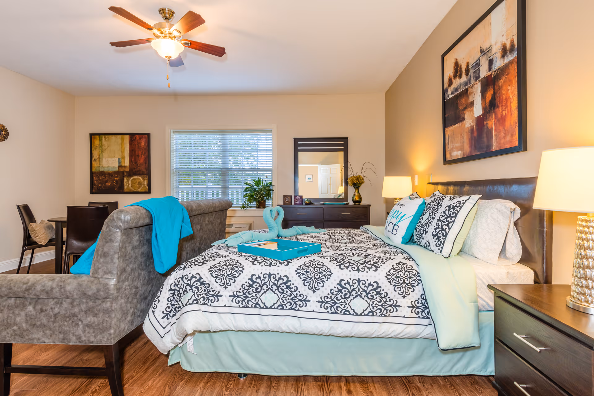 A well-lit bedroom featuring a bed with patterned bedding and decorative pillows, a nightstand with a lamp, a dresser with a mirror and plants, a ceiling fan with light, a gray upholstered chair with a turquoise throw, and a small dining table with chairs near a window with blinds.