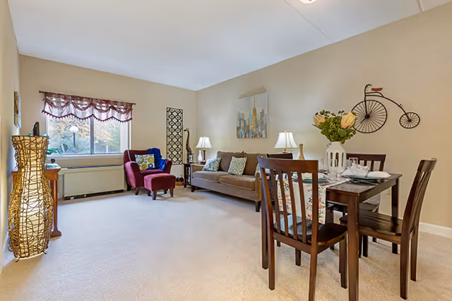 A cozy living and dining area with beige walls and carpet. The living area features a brown sofa with decorative pillows, a red armchair with a blue pillow, two table lamps on side tables, and a window with a valance. The dining area has a dark wooden table set for four with a floral table runner and a vase of flowers. Wall decorations include a cityscape painting and a vintage bicycle sculpture.