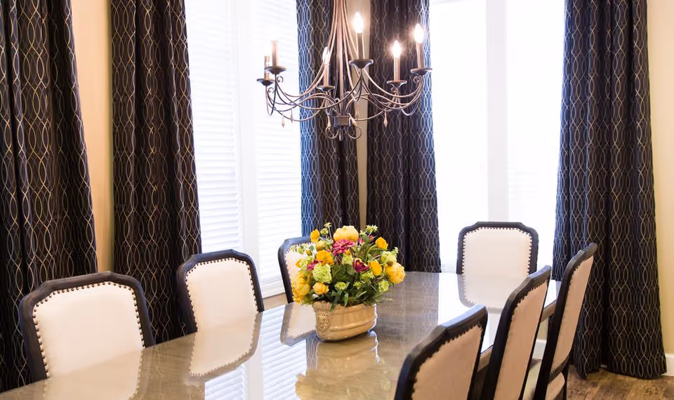 Dining room with a glass-topped table surrounded by upholstered chairs, a floral centerpiece, a chandelier, and tall windows with patterned curtains.