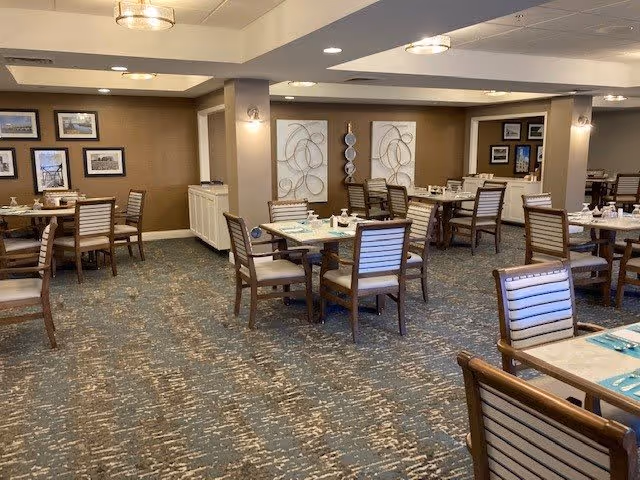 A spacious dining room with multiple wooden tables and chairs arranged neatly. The tables are set with placemats, napkins, and small condiment containers. The room has soft lighting with ceiling fixtures and wall sconces, neutral-colored walls adorned with framed pictures and decorative wall art. The carpet has a patterned design in shades of blue and beige.