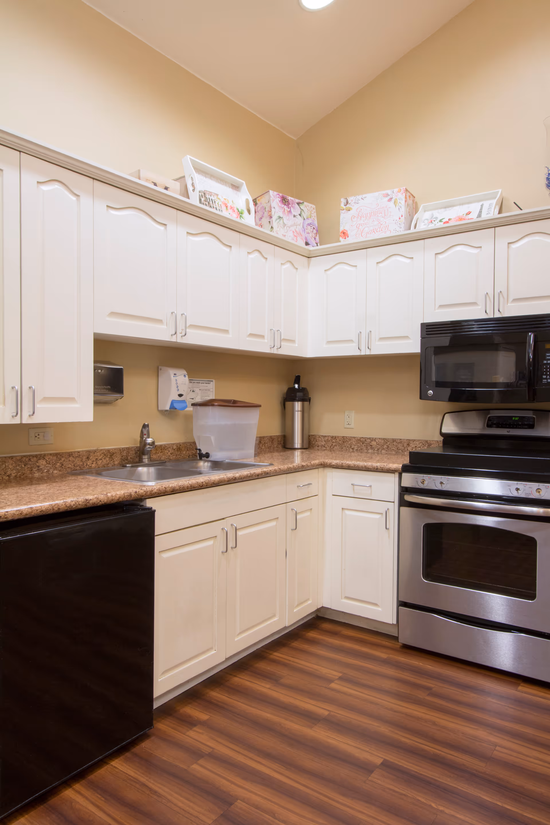 A kitchen area with white cabinets, a stainless steel oven and microwave, a black mini refrigerator, a double sink, and a countertop with a water dispenser and a coffee dispenser. The floor is wooden and the walls are beige. Decorative boxes and framed pictures are placed on top of the cabinets.