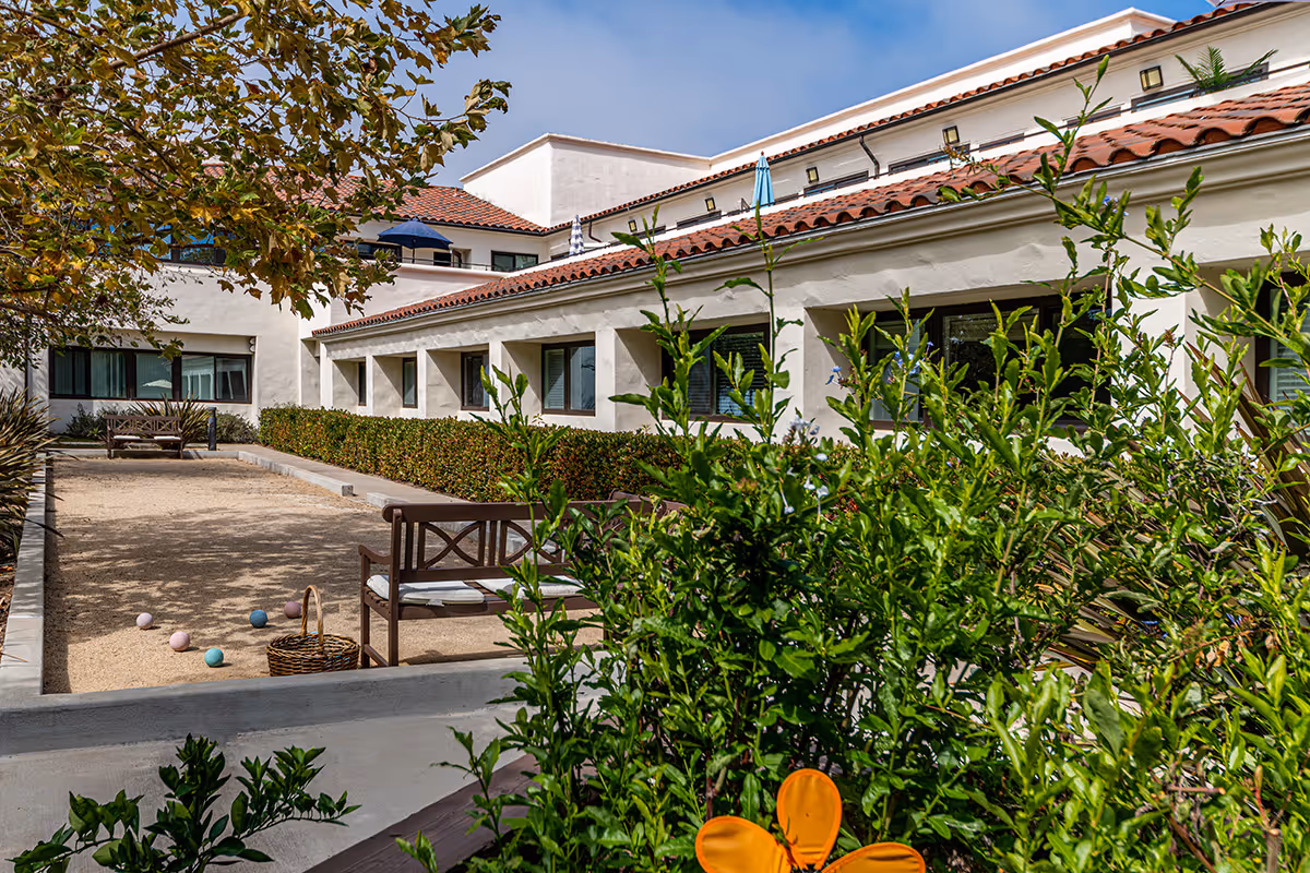 Outdoor courtyard area at GranVida Senior Living and Memory Care featuring a bocce ball court with several bocce balls and a basket, wooden benches with cushions, surrounded by green bushes and trees, with a white building with red-tiled roof in the background under a clear blue sky.