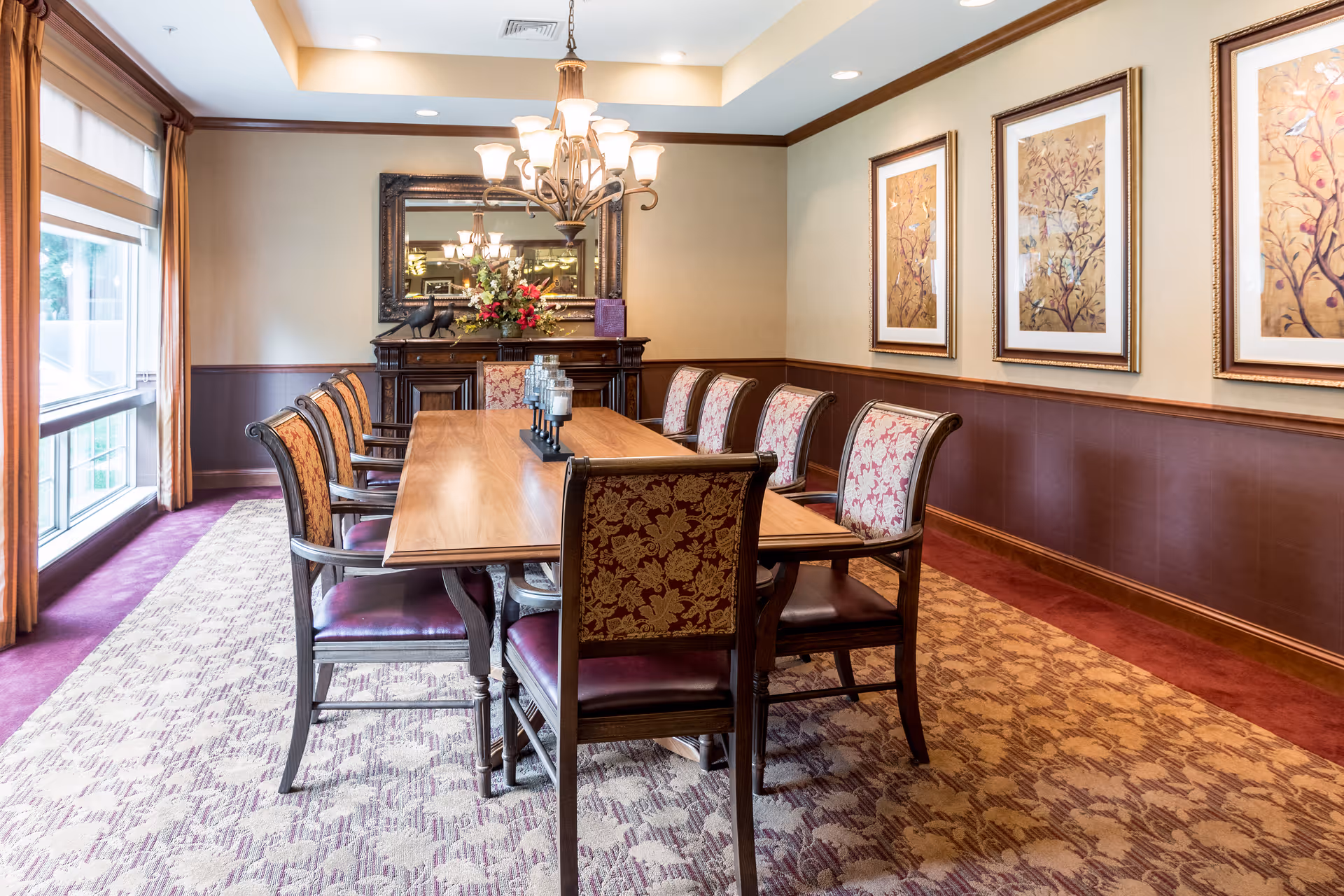 A well-lit dining room with a long wooden table surrounded by eight upholstered chairs with floral patterns. The room features a large window with curtains on the left, a chandelier hanging above the table, a decorative sideboard with a mirror and flower arrangement at the far end, and three framed floral artworks on the right wall. The carpet has a patterned design in shades of beige and purple.