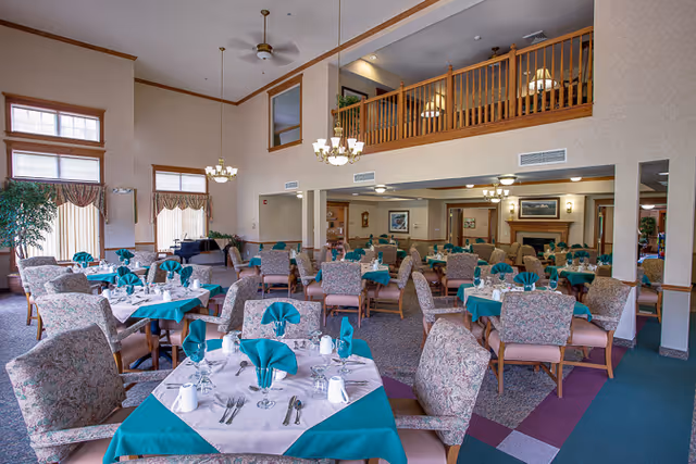 A spacious dining room with multiple tables covered in white and teal tablecloths, each set with teal folded napkins, silverware, glasses, and white mugs. The room features high ceilings with ceiling fans and chandeliers, large windows with curtains, and a second-floor balcony overlooking the area. Comfortable upholstered chairs surround the tables, and there is a fireplace and framed artwork on the walls in the background.
