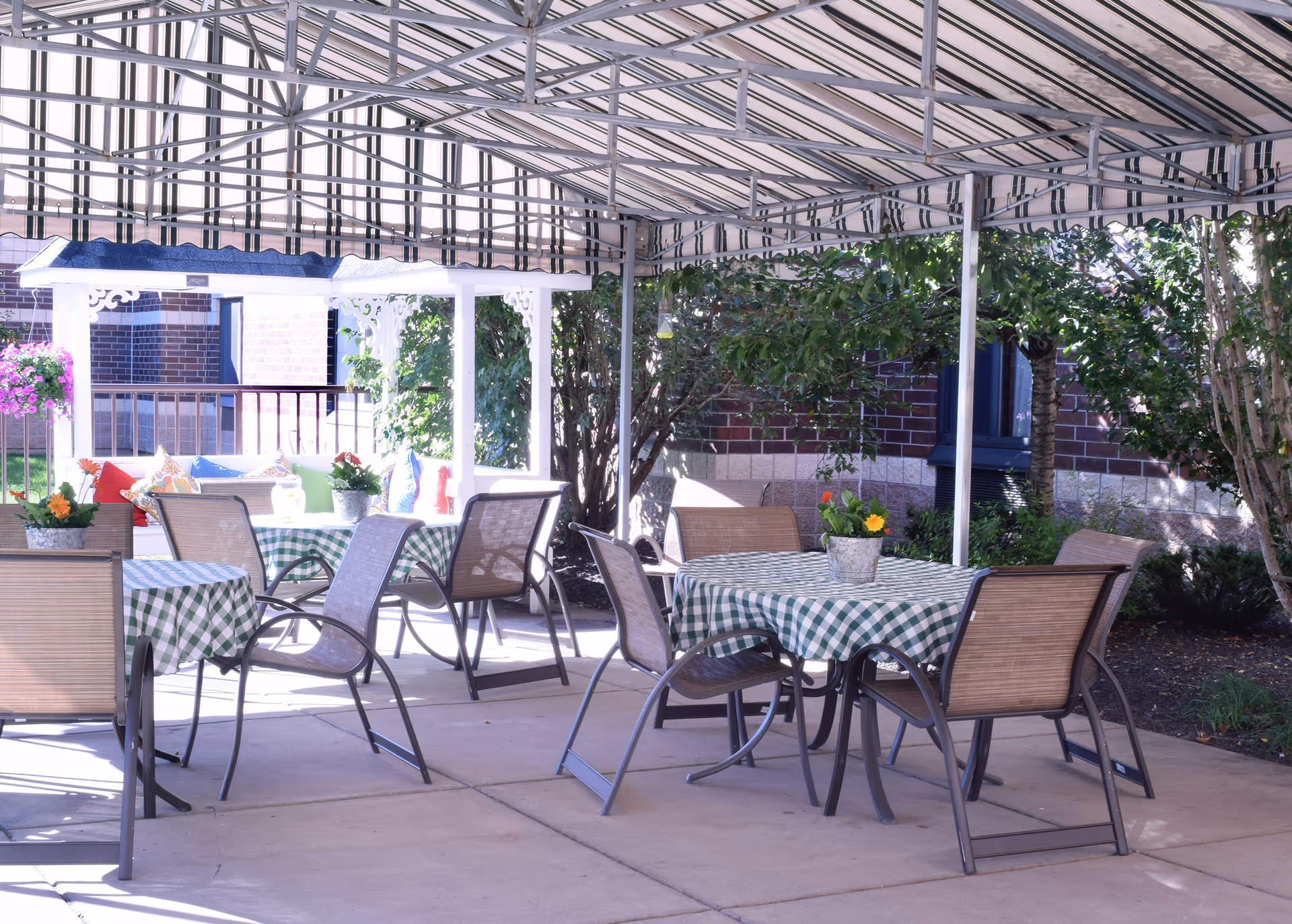 Outdoor patio area with several tables covered in green and white checkered tablecloths, surrounded by chairs. The space is shaded by a striped canopy and decorated with potted flowers on the tables. There are trees and bushes around the patio, and a brick building is visible in the background.