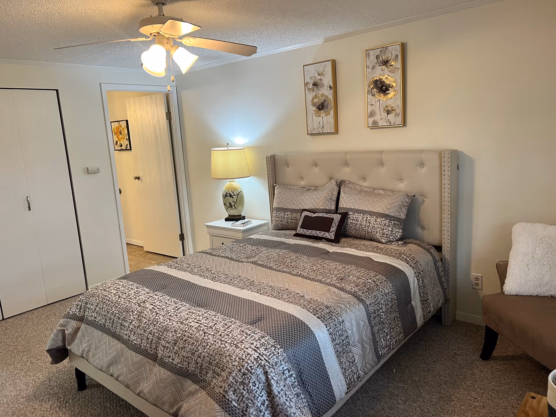 A neatly made bed with a patterned gray and beige comforter and pillows in a bedroom. There is a beige upholstered headboard with button tufting. On the right side of the bed is a white nightstand with a decorative lamp. Two floral paintings hang on the wall above the bed. A ceiling fan with lights is on the ceiling. A brown chair with a white pillow is partially visible on the right. A closet with white doors and an open doorway leading to another room are visible on the left.