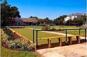 Outdoor bocce ball courts with green playing surfaces, surrounded by landscaped plants and trees, under a clear blue sky. Residential buildings are visible in the background.