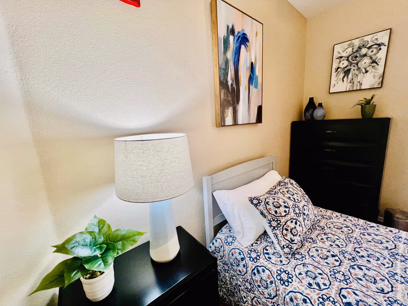 A cozy bedroom with a single bed featuring a patterned blue and white bedspread and matching pillow. Next to the bed is a black nightstand with a white table lamp and a small green potted plant. On the wall above the bed are two pieces of artwork, one abstract and one floral. In the corner, there is a black chest of drawers with decorative vases and another small plant on top.