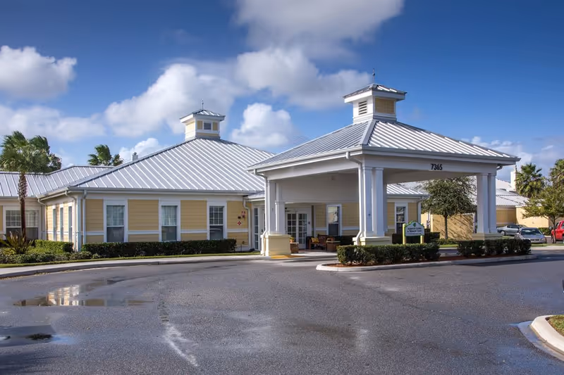Exterior view of The Brennity at Melbourne Senior Living building with a covered entrance, yellow walls, white trim, and a metal roof under a partly cloudy sky.