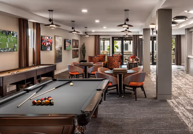 A spacious game and lounge room with a pool table in the foreground, a shuffleboard table along the left wall, and several round tables with chairs. The room has multiple ceiling fans, wall-mounted televisions showing sports, and a seating area with orange and patterned cushions near large windows and glass doors leading outside.