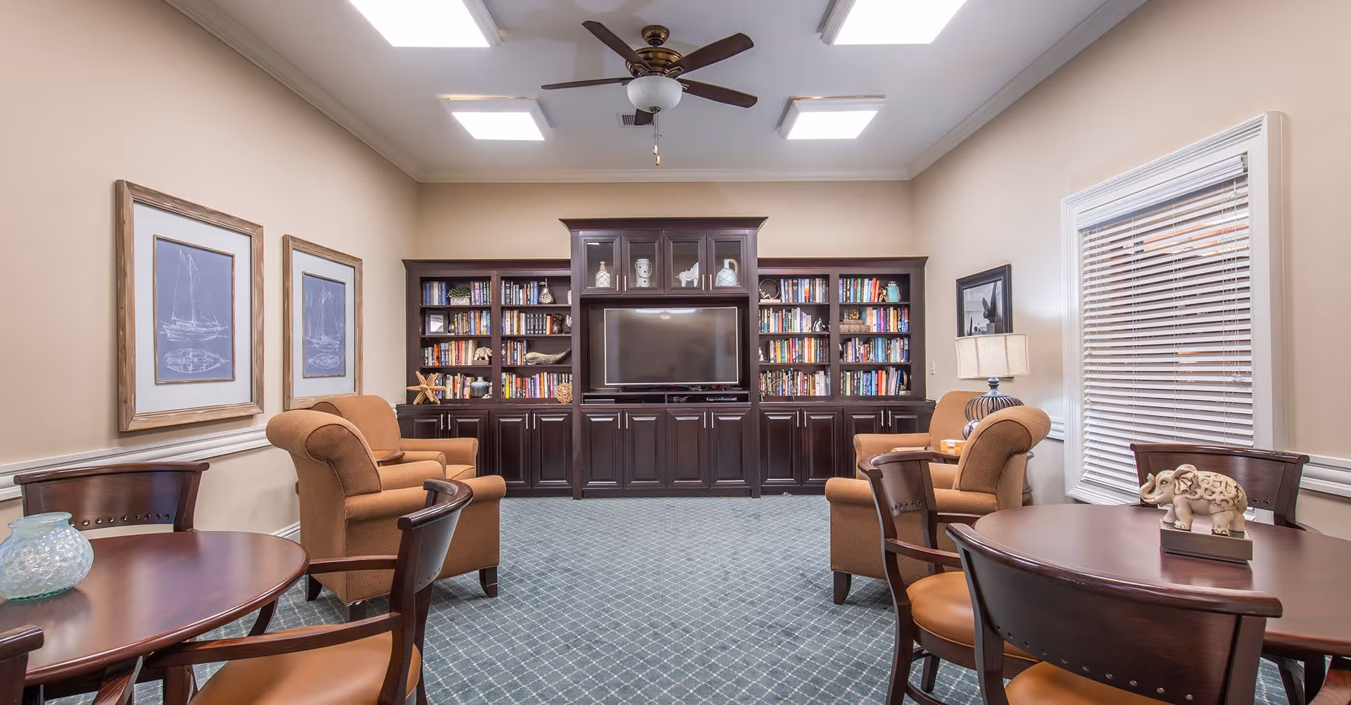 A cozy living room with beige walls and a blue patterned carpet. The room features a large dark wood entertainment center with a flat-screen TV and bookshelves filled with books and decorative items. There are four comfortable brown armchairs arranged around the room, two on each side. Two round wooden tables with chairs are positioned in the foreground, each table decorated with small ornaments. The room is well-lit with ceiling lights and a ceiling fan, and there are framed nautical-themed pictures on the left wall and a window with blinds on the right.