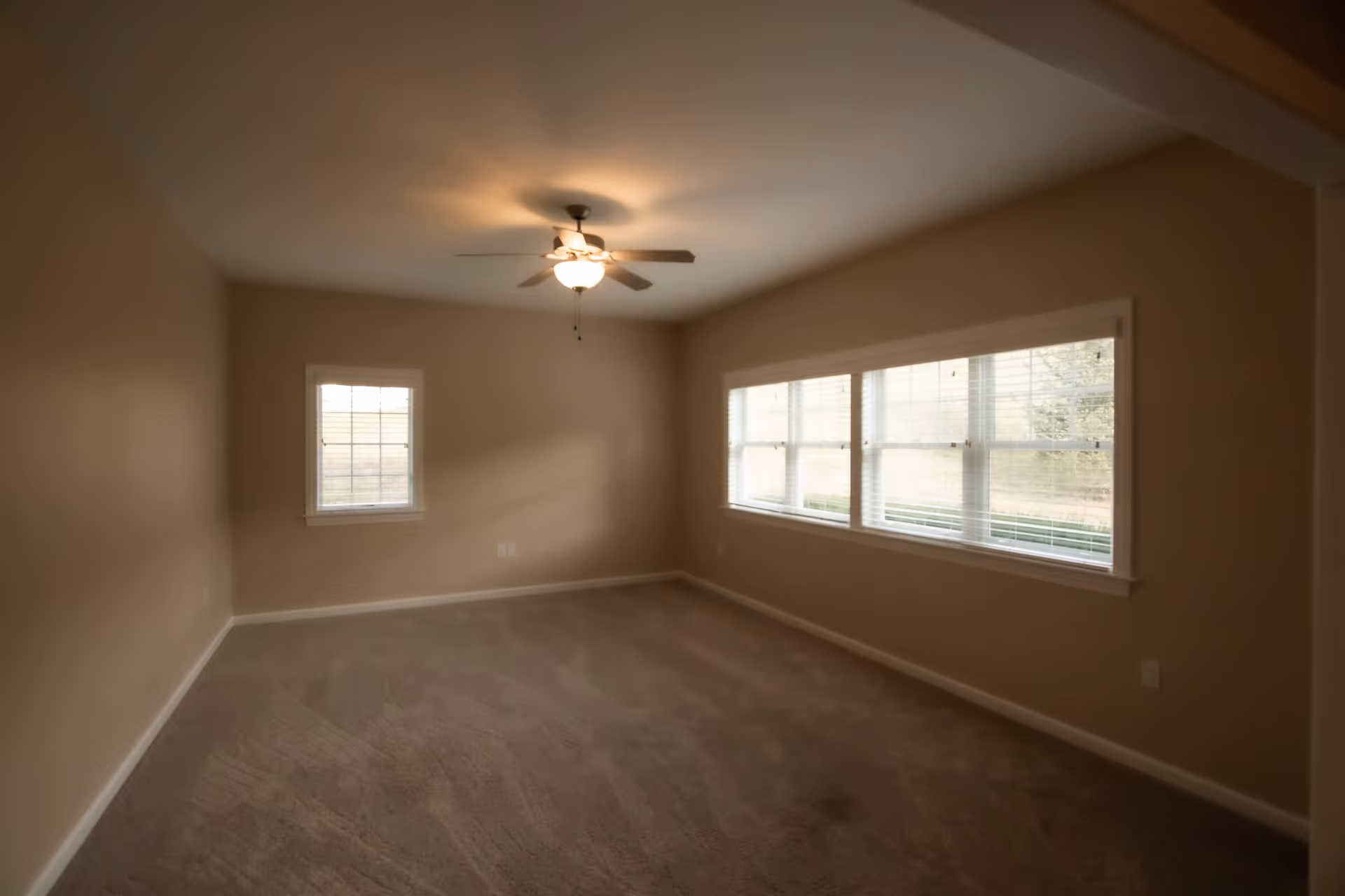 Empty room with beige walls and carpeted floor, featuring a ceiling fan with light fixture and multiple windows with blinds allowing natural light to enter.