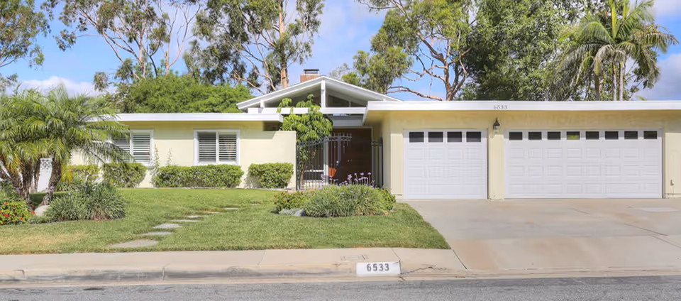 Front exterior view of a single-story residential building with a well-maintained lawn, shrubs, palm trees, and a driveway leading to two white garage doors under a clear blue sky.