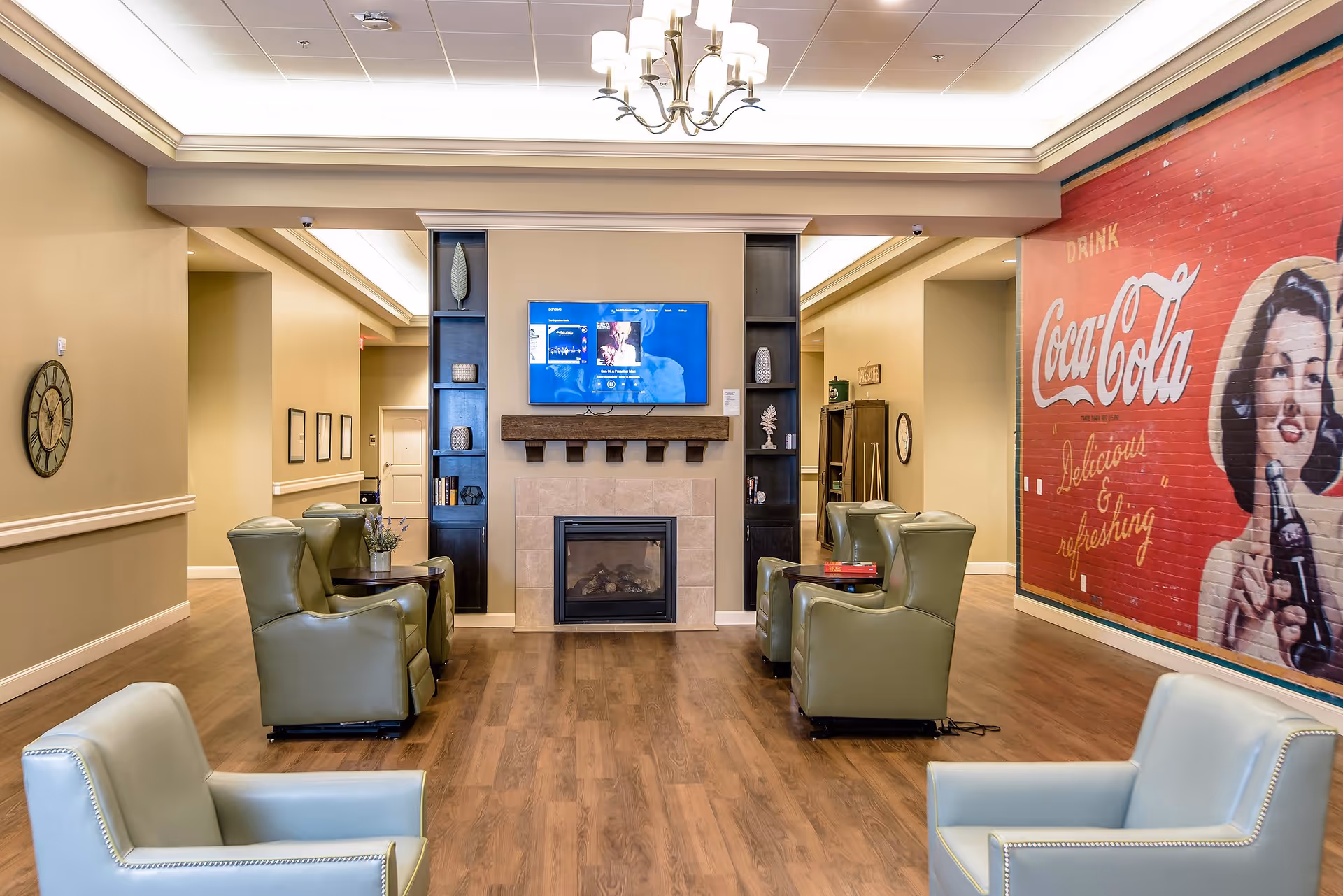 A cozy common area with six green armchairs arranged around a fireplace with a mounted flat-screen TV above it. The room has wooden flooring, beige walls, and a large vintage Coca-Cola mural on the right wall. Decorative shelves flank the fireplace, and a clock is mounted on the left wall.