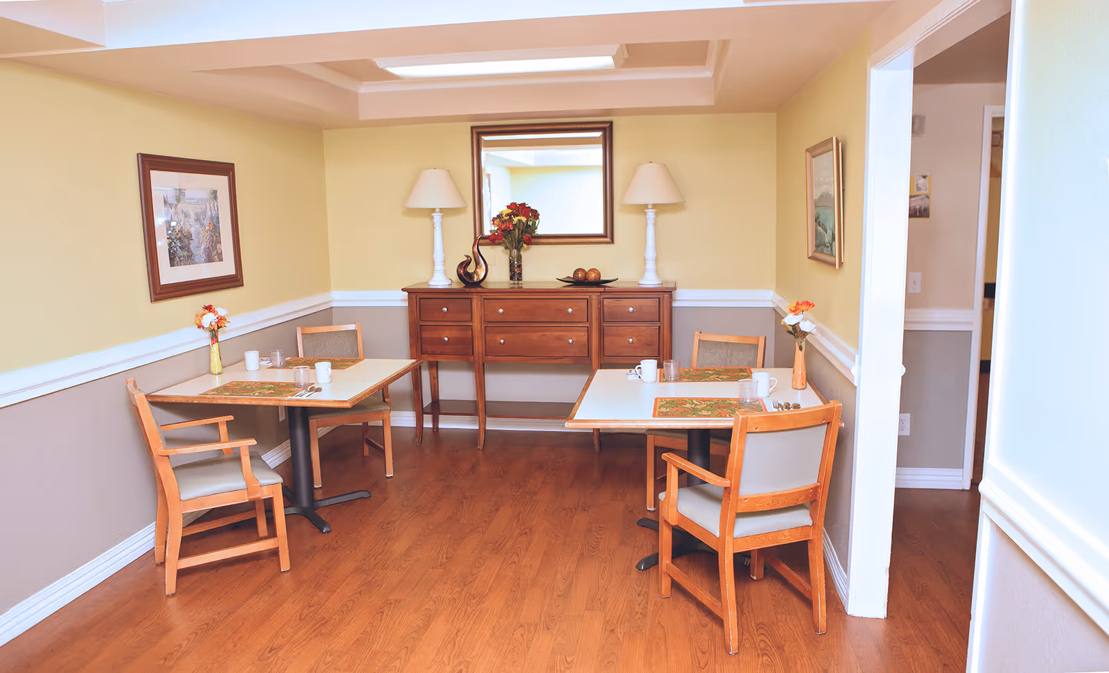 Cozy dining room with two small tables and wooden chairs, a wooden sideboard with lamps and a mirror, and wood floors.