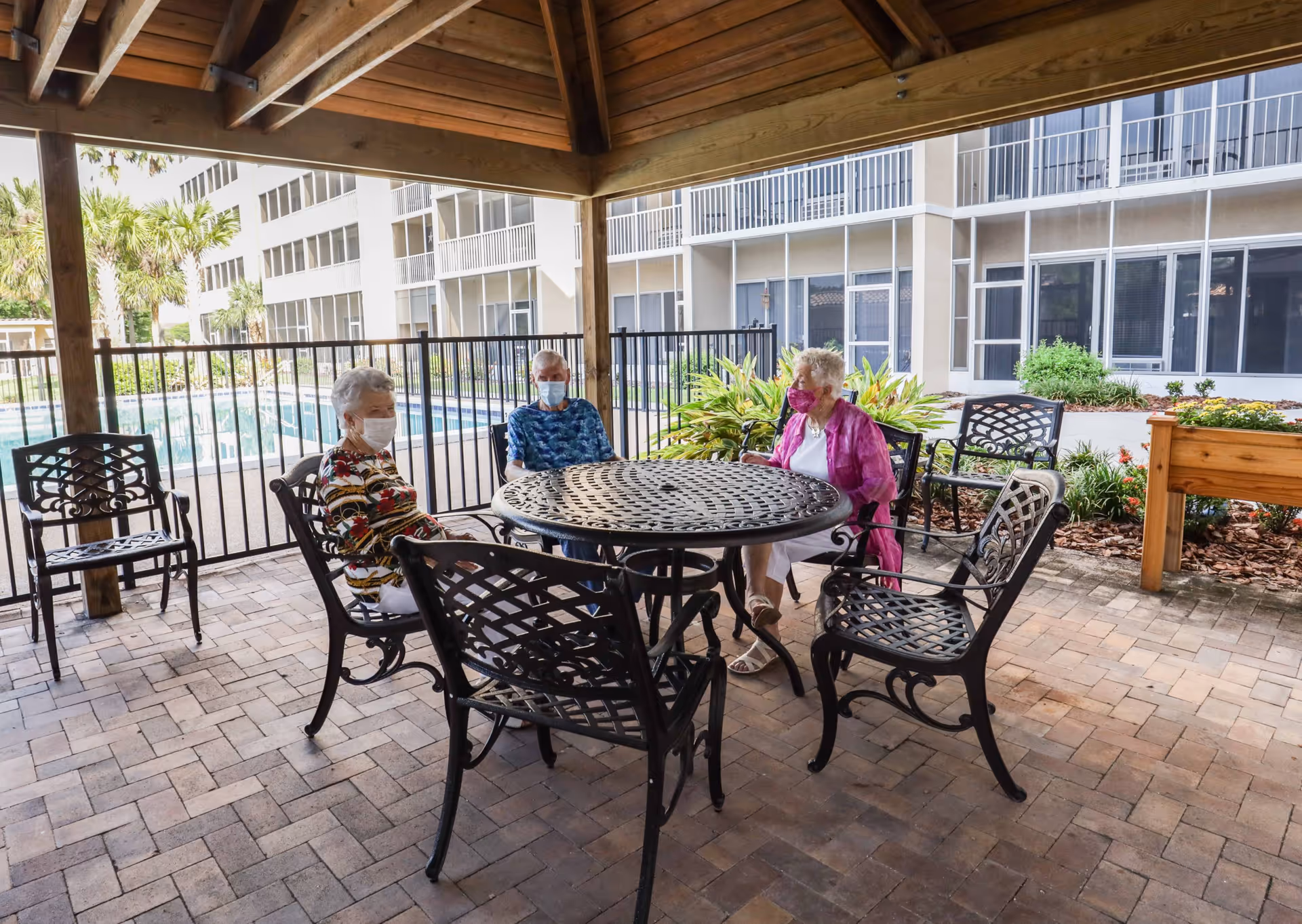 Three elderly women wearing face masks sitting around a round metal table under a wooden gazebo near a swimming pool in an outdoor area of a retirement community. The background shows a multi-story building with balconies and windows, along with some greenery and plants.