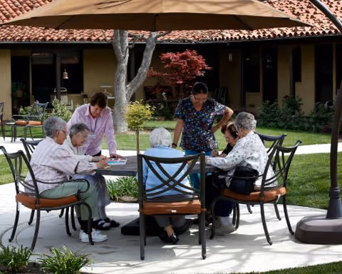 A group of elderly women sitting around a round outdoor table under a large umbrella, engaged in an activity with two caregivers standing nearby. The setting is a courtyard with green grass, plants, and a building with a tiled roof in the background.