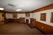 Interior view of a common area with brown carpet, wood-paneled lower walls, and white upper walls. The room features built-in wooden shelves with books and a small fireplace. There are framed paintings on the walls and a flat-screen TV mounted above the fireplace. The ceiling has recessed lighting and an air vent.