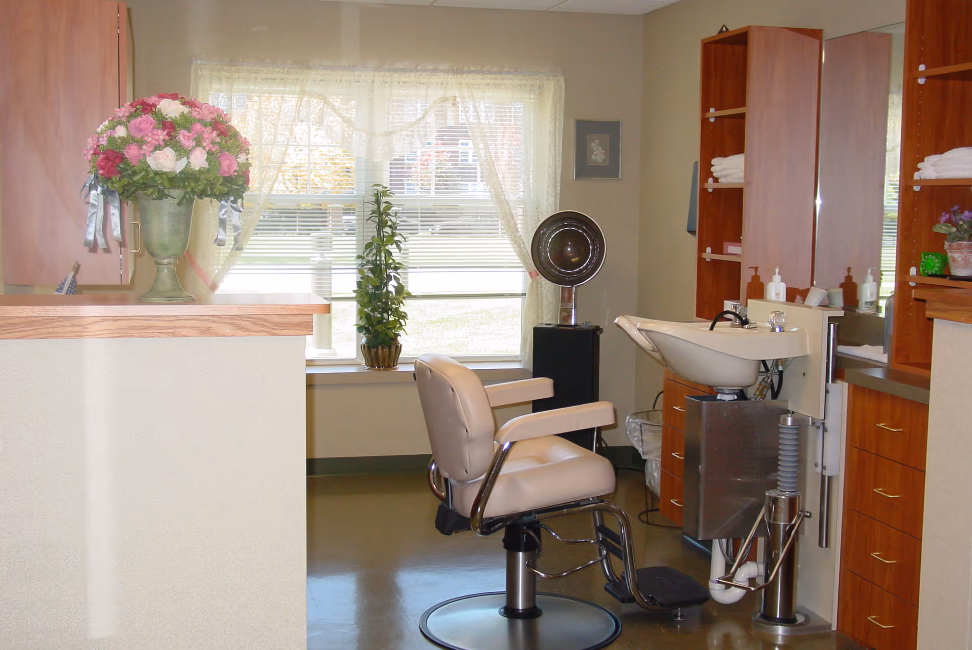 Interior of a hair salon area in a senior living facility with a salon chair, a hair washing sink, wooden cabinets with towels and hair products, a large window with lace curtains, and a vase of pink and white flowers on a counter.