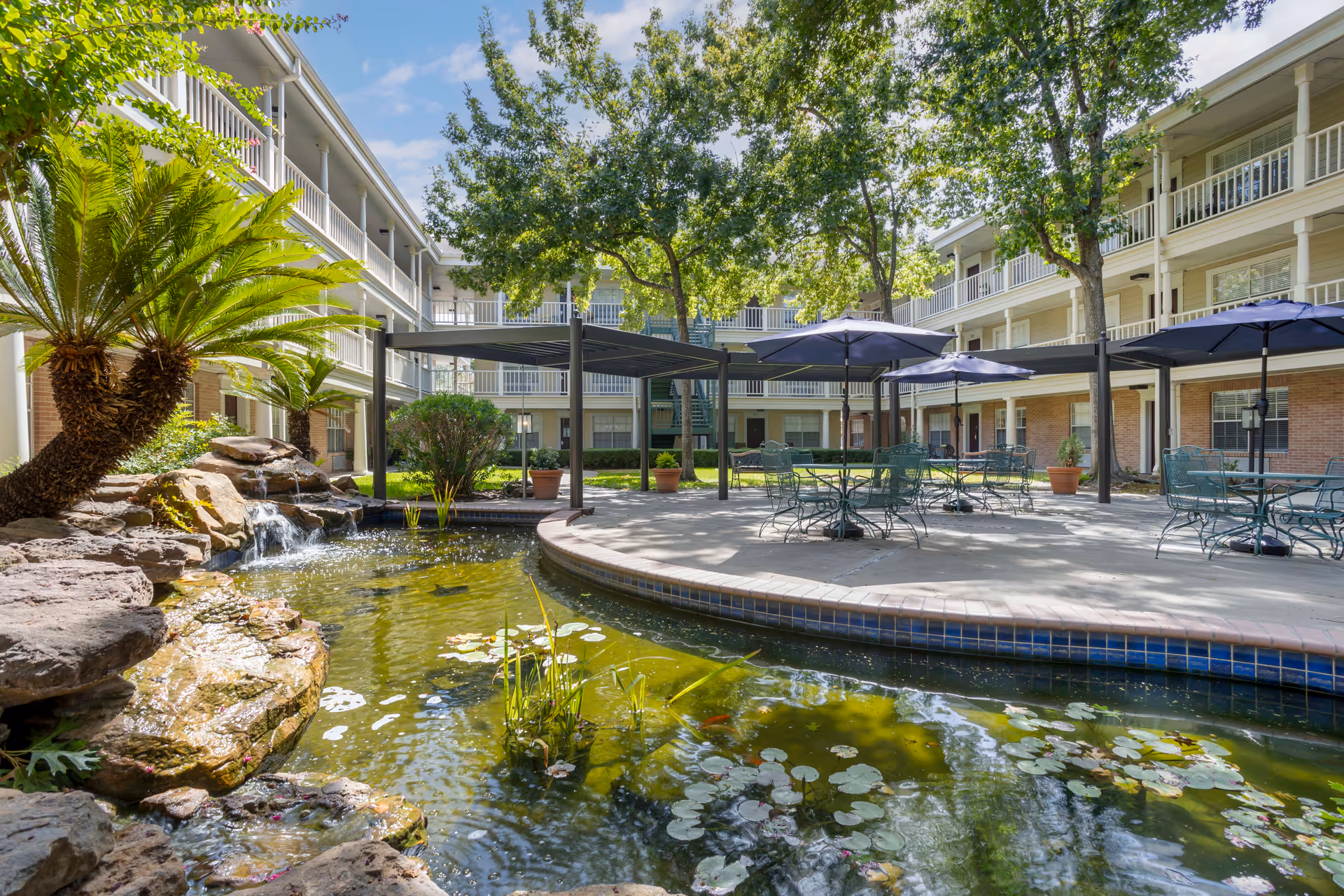 Sunny courtyard with a koi pond and small waterfall, patio tables and umbrellas, and surrounding three-story balcony walkways.