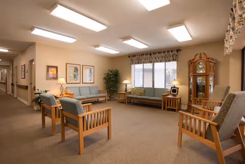 A well-lit living room area in a senior living facility featuring wooden framed chairs and sofas with cushions, a grandfather clock, side tables with lamps, framed artwork on the walls, a large window with curtains, and a potted plant in the corner.