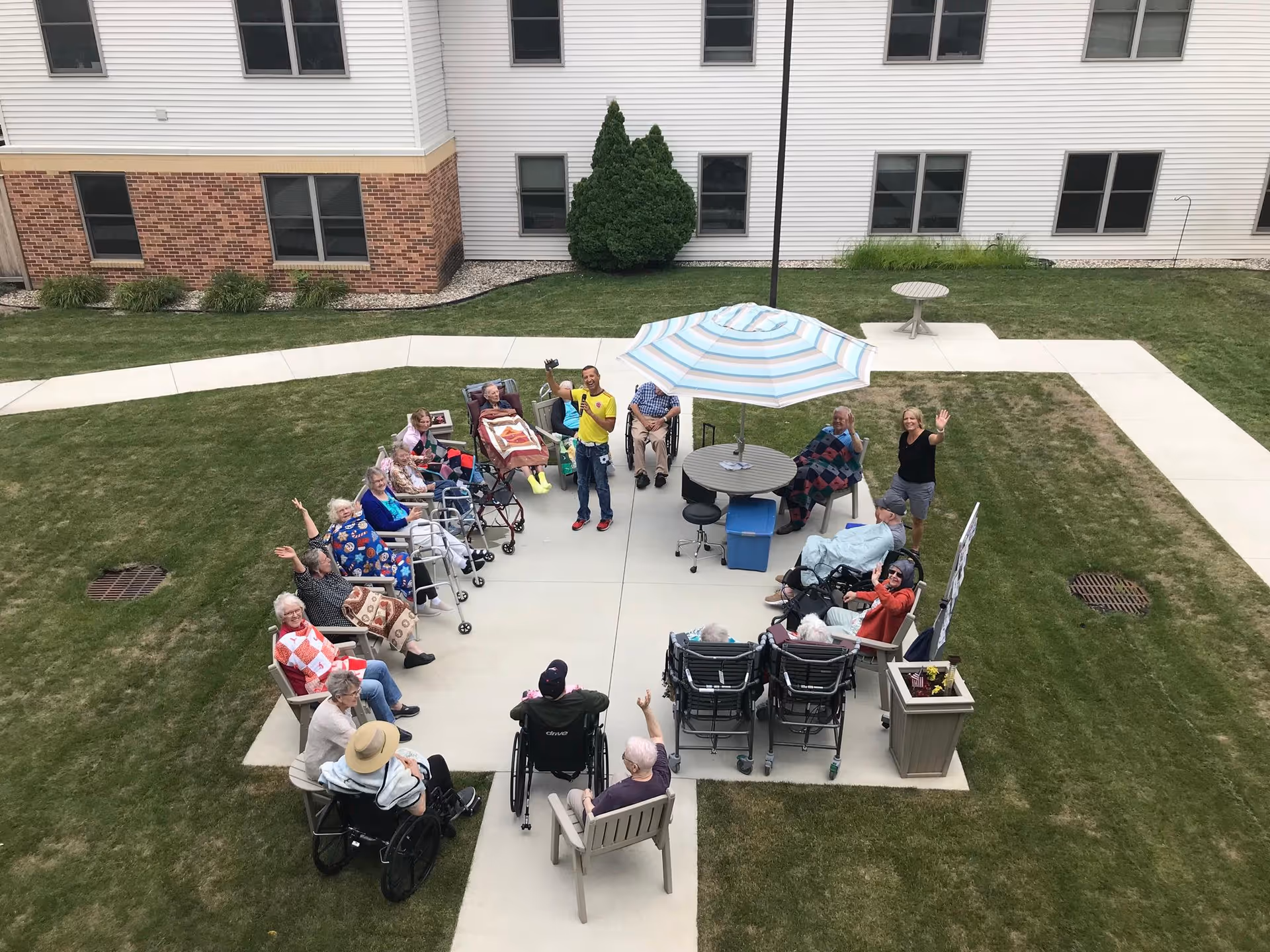 A group of elderly people sitting in a circle outdoors on a concrete patio area, some in wheelchairs and some in chairs, with a few staff members standing and waving. There is a round table with a blue and white striped umbrella in the center, surrounded by grass and a building with windows in the background.