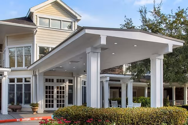 Entrance of a senior living facility with a covered drop-off area supported by white columns, surrounded by bushes and trees. The building has large windows and a beige exterior with white trim.