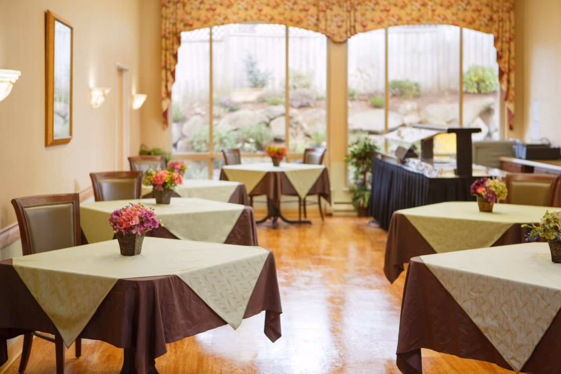 A dining room with several square tables covered with brown and beige tablecloths, each table decorated with a small flower arrangement. The room has large windows with floral valances, letting in natural light and showing a garden area outside. Chairs are placed around the tables, and there is a buffet station along one wall.