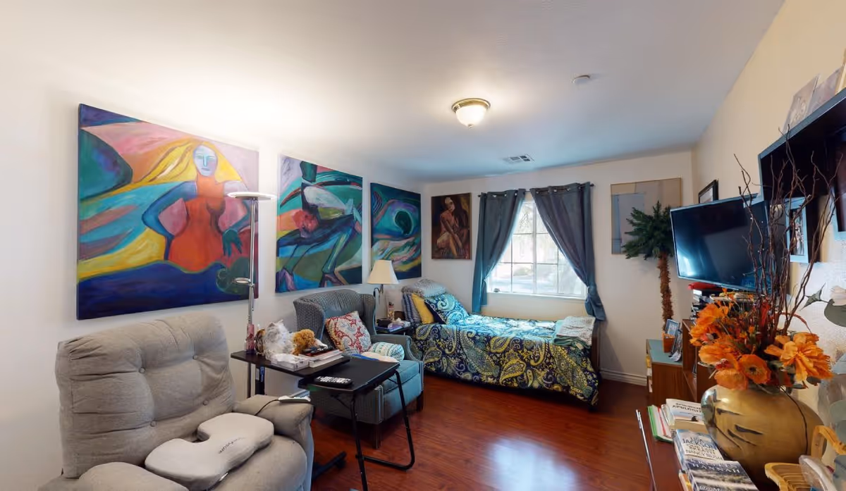 A cozy bedroom in a senior living facility featuring a single bed with a colorful paisley bedspread, a window with dark curtains, two armchairs with a small table between them, vibrant abstract paintings on the wall, a flat-screen TV mounted on the wall, and a vase with orange flowers on a cabinet.