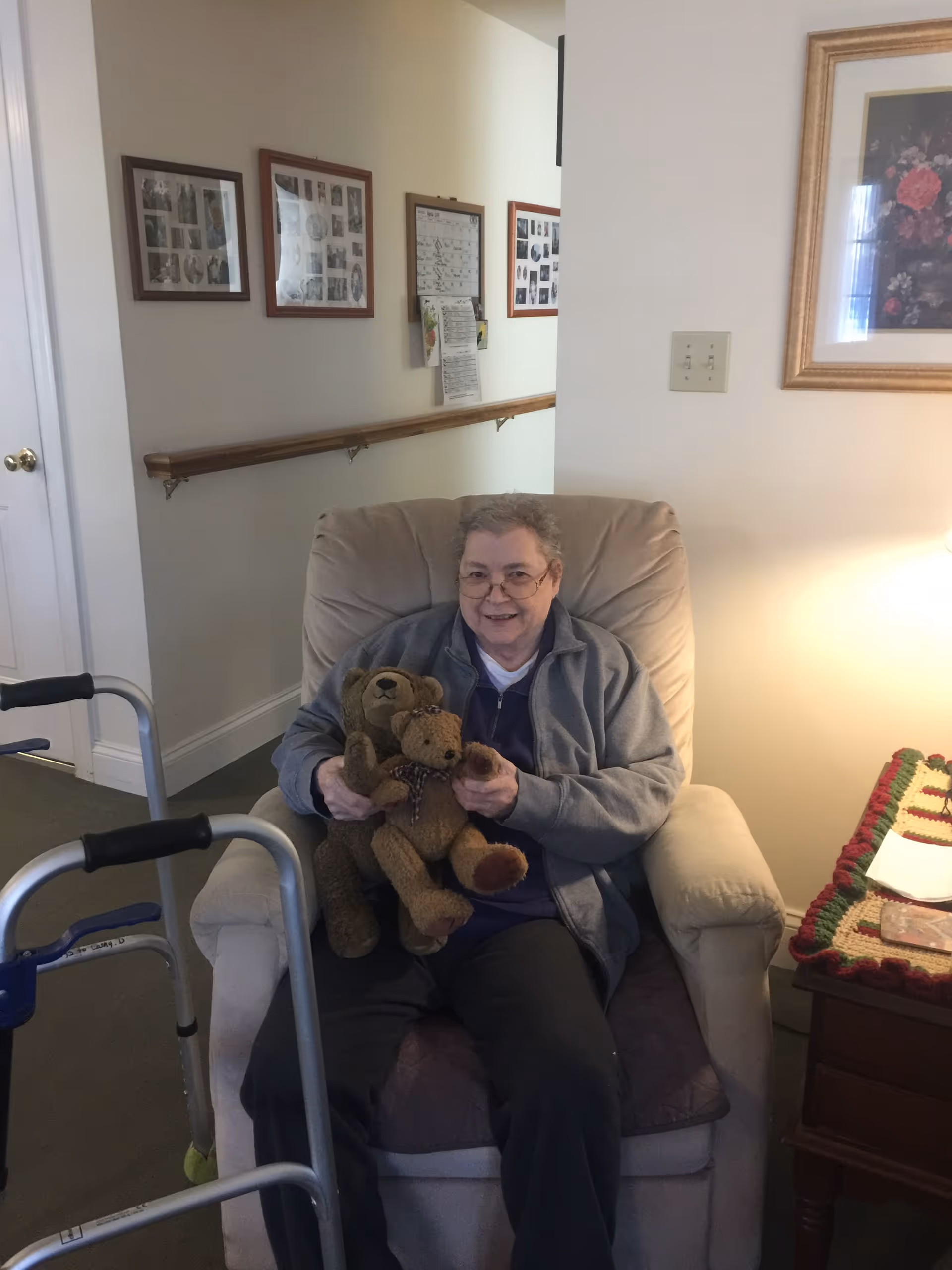 An older woman sitting in a chair holding two stuffed bears in a cozy room.