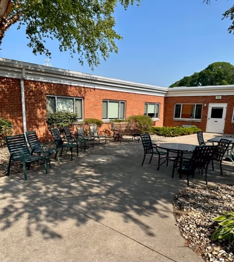 Outdoor patio area at a senior living facility with several green metal chairs arranged along a brick building wall and around a round table. There are bushes and small plants along the building, and a tree casting shadows on the concrete ground under a clear blue sky.