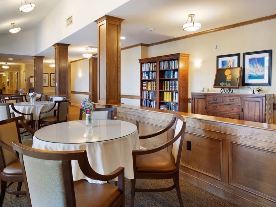 A well-lit common area with round tables covered in white tablecloths and surrounded by wooden chairs with cushioned seats. Small flower arrangements in glass jars are placed on the tables. The room features wooden columns, a wooden bookshelf filled with books, and a wooden sideboard with framed artwork and decorative items on top. The walls are light-colored with wood trim, and ceiling lights provide warm illumination.