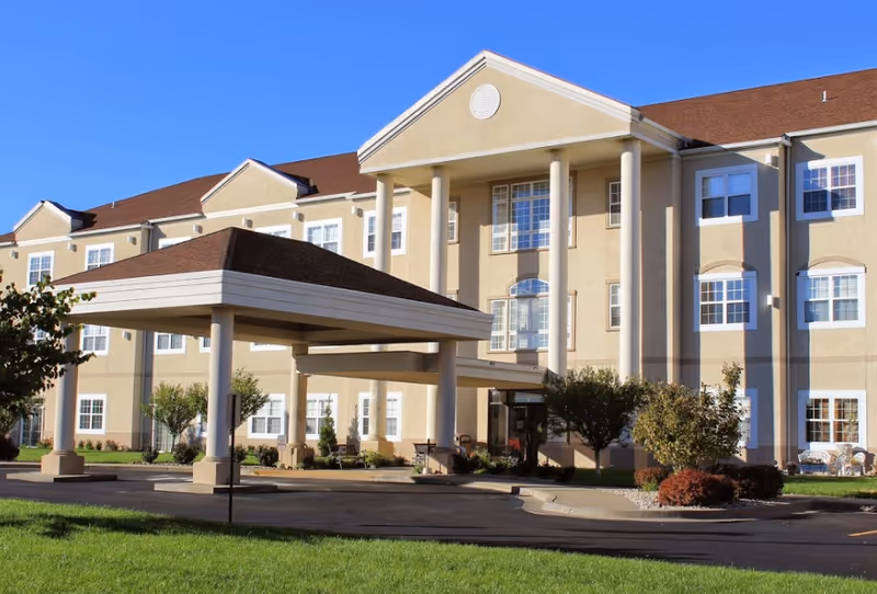 Exterior view of a multi-story beige retirement community building with white columns and a covered entrance. The building has multiple windows and is surrounded by a well-maintained lawn and small trees under a clear blue sky.