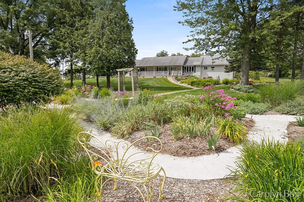 Landscaped garden with winding paths, flower beds and a decorative bench in front of a single-story building surrounded by trees.