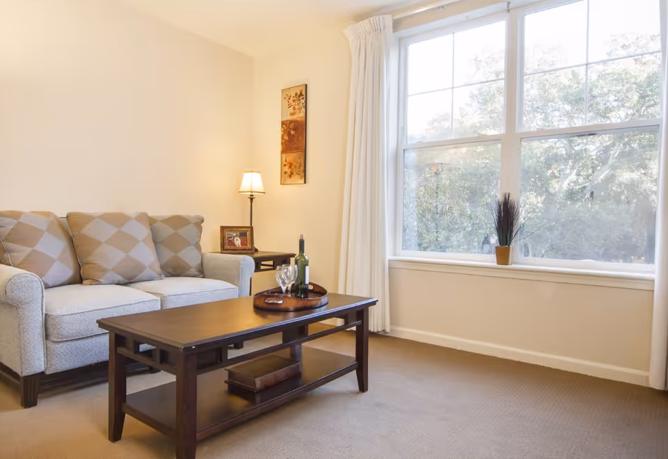 A cozy living room with a beige sofa adorned with three patterned cushions, a wooden coffee table with a tray holding a bottle of wine and two glasses, a side table with a lamp and a framed photo, and a large window with white curtains letting in natural light and showing trees outside.