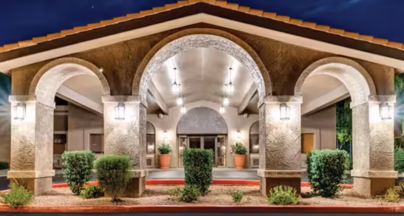 The front entrance of Springdale Village Assisted Living Facility & Independent Living Apartments in Mesa, Arizona, featuring a covered driveway with three large stone arches, outdoor lighting fixtures, and neatly trimmed bushes in front of the building at dusk.