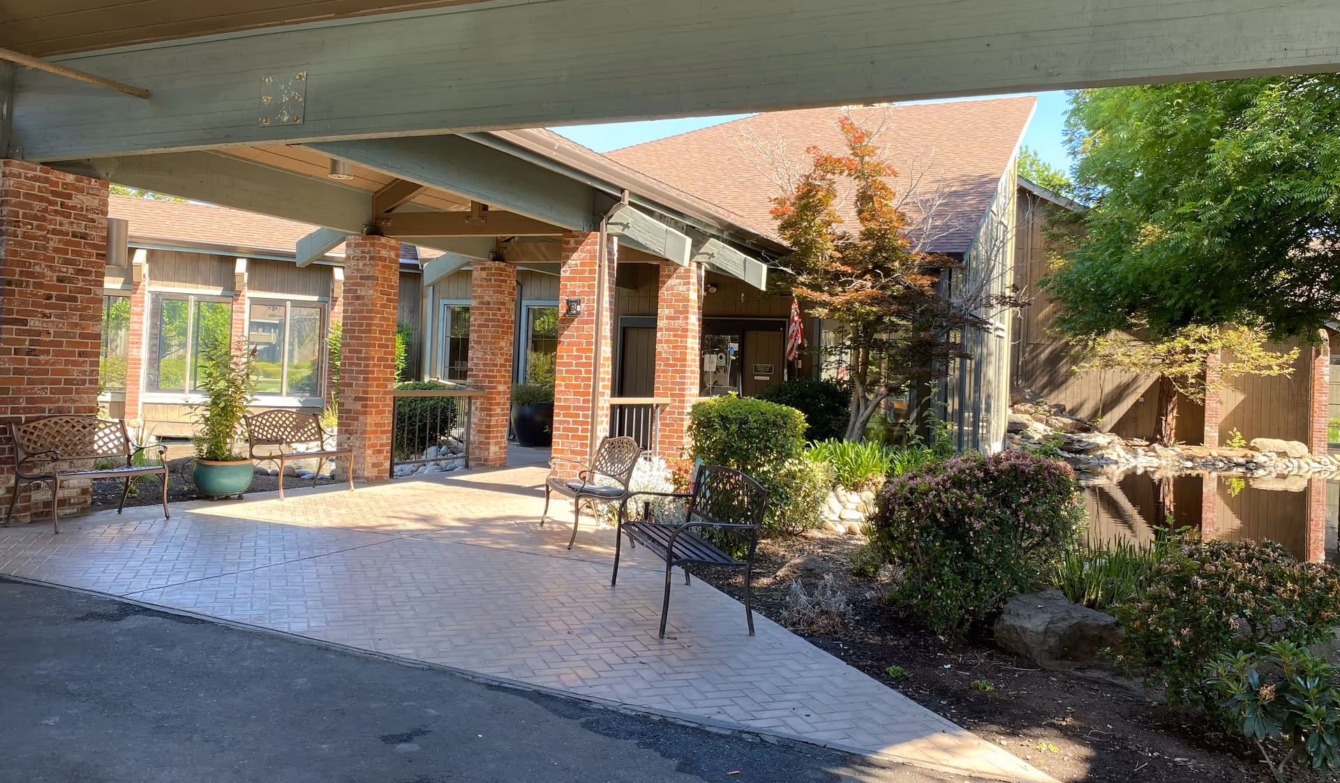 Covered entrance to a brick senior living building with benches, landscaping and a reflecting pond.
