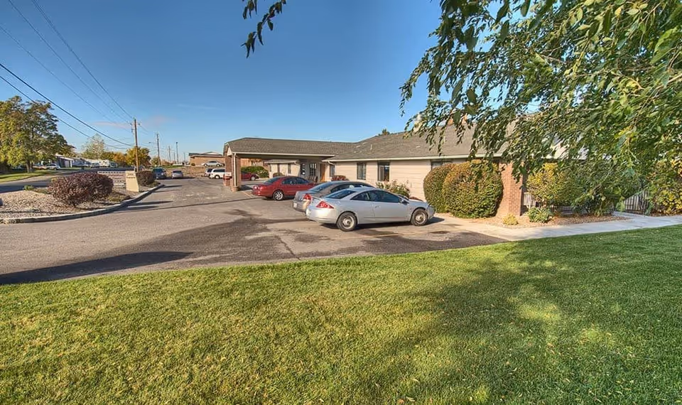 Exterior view of Logan Creek Retirement Community showing a single-story building with a parking lot in front. Several cars are parked, and there is a well-maintained grassy area with trees and bushes surrounding the building under a clear blue sky.