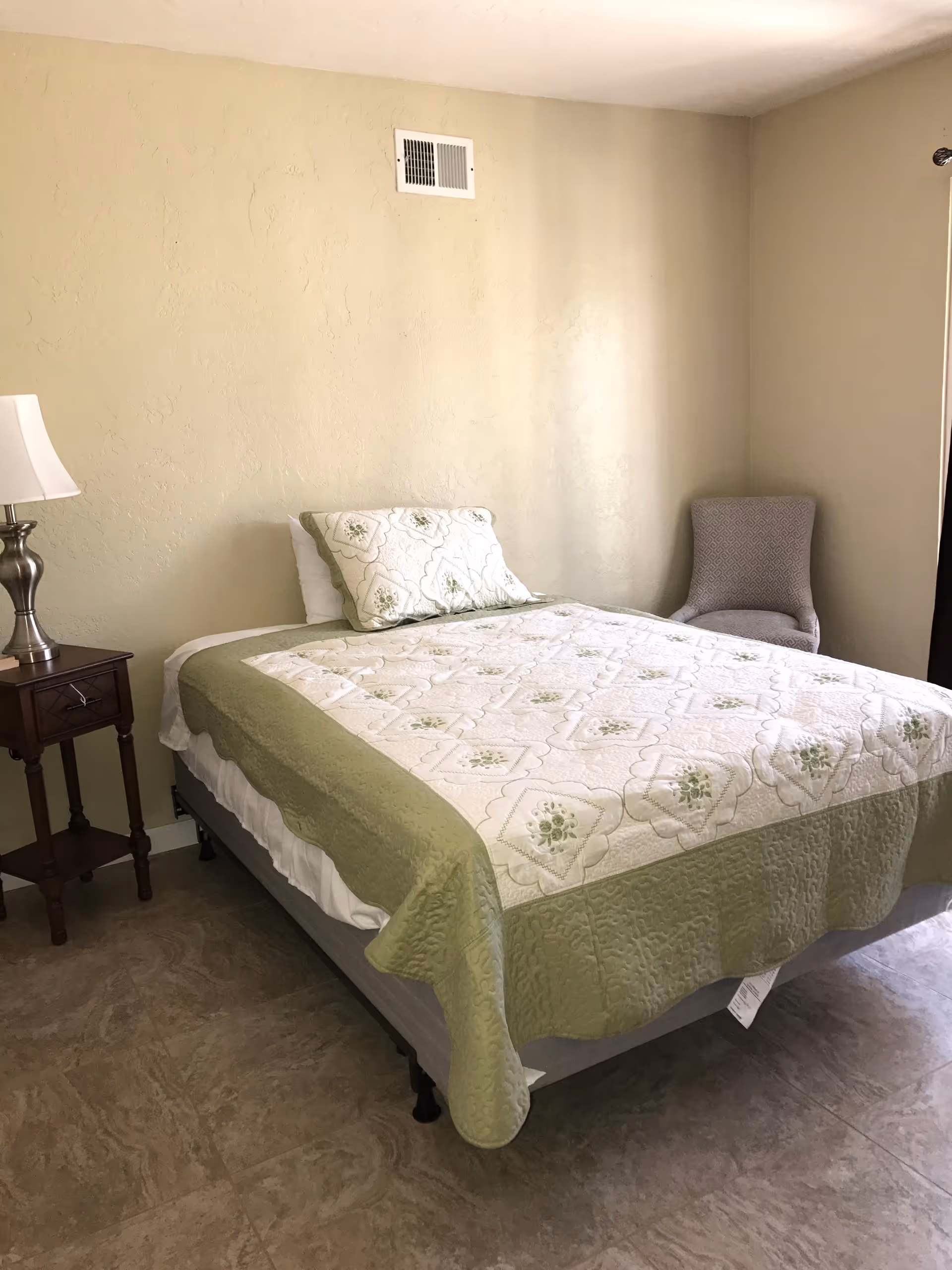 A simple bedroom with a bed covered in a white and green quilt, a matching pillow, a wooden nightstand with a lamp, and a gray upholstered chair in the corner near a window with dark curtains.