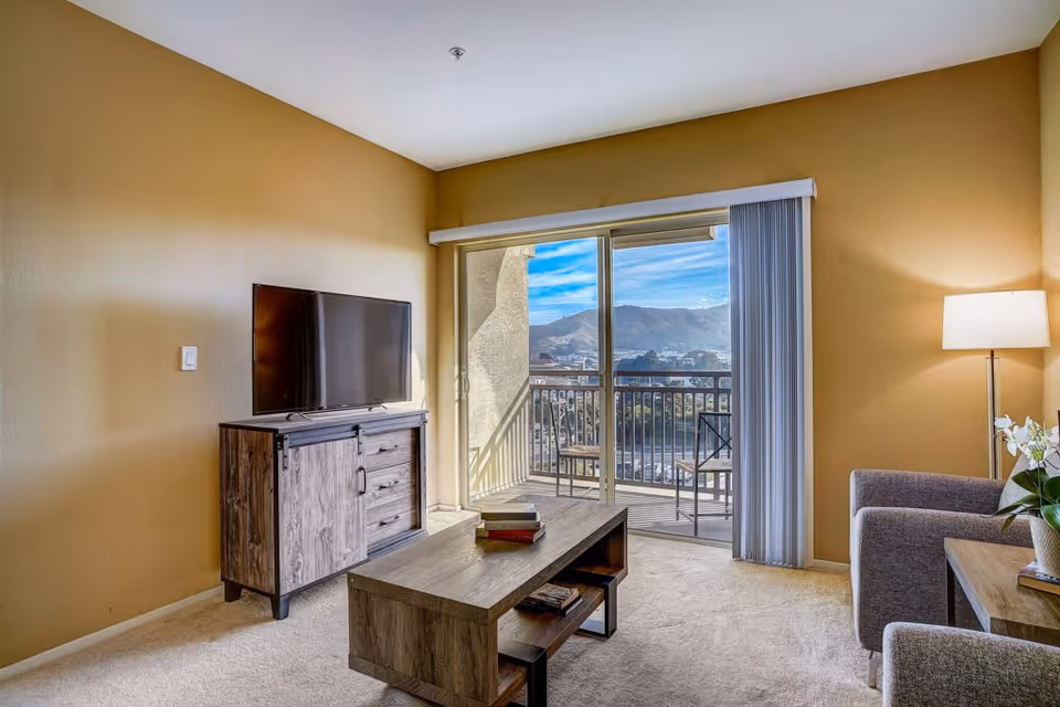 A cozy living room with beige walls and carpet, featuring a flat-screen TV on a wooden cabinet, a wooden coffee table with books, a gray armchair, a floor lamp, and a sliding glass door leading to a balcony with a view of mountains and a clear blue sky.
