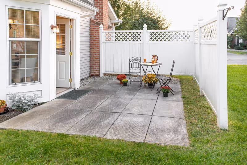 A small outdoor patio area with a concrete floor, a white privacy fence, and a brick and white house exterior. There is a small round table with two metal chairs, a pitcher and glasses on the table, and several potted flowers placed around the patio.