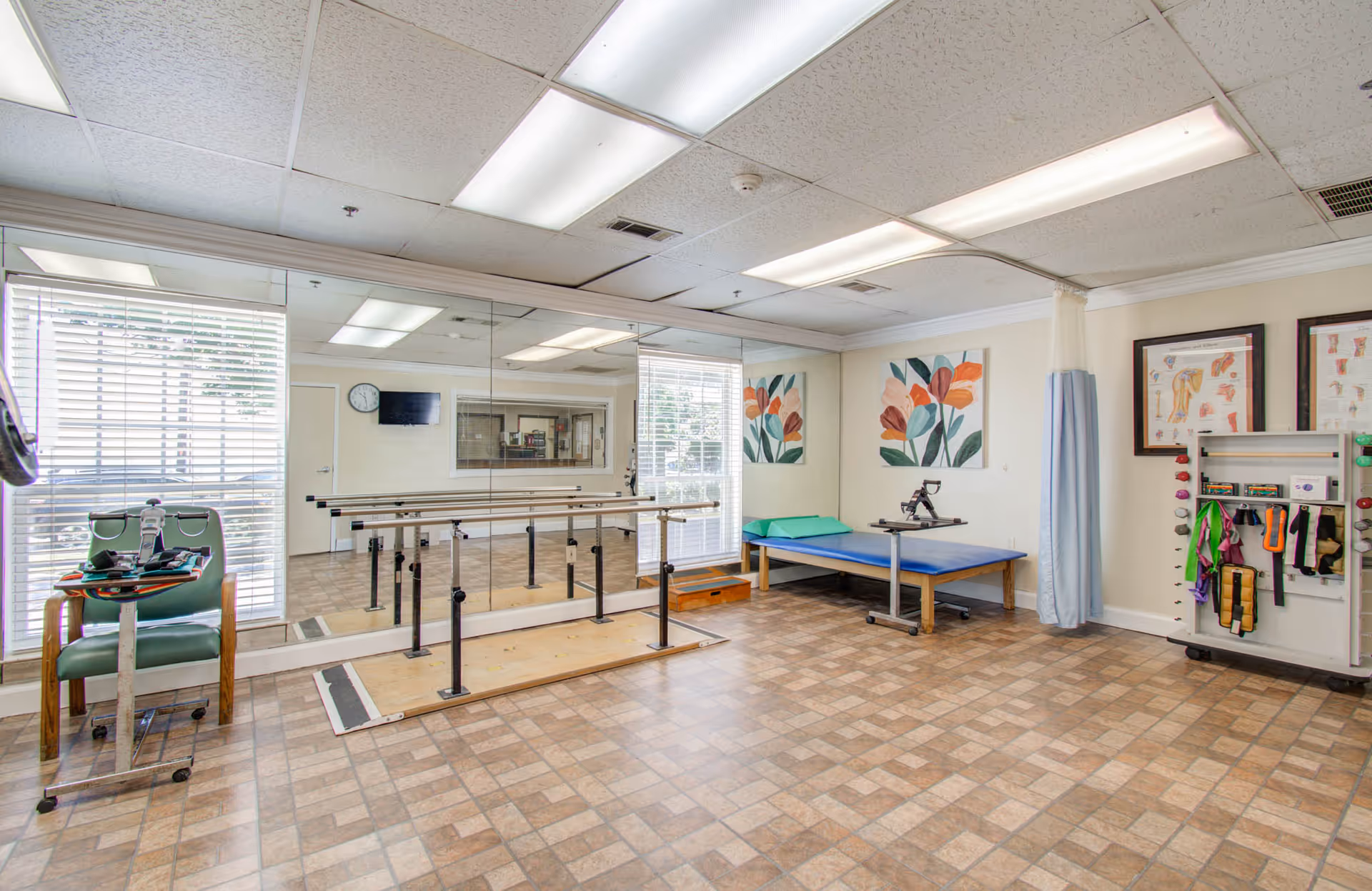 A physical therapy room with parallel bars for walking exercises, a green chair with exercise equipment, a blue therapy table with a green pillow, anatomical charts on the walls, and large windows with blinds allowing natural light.